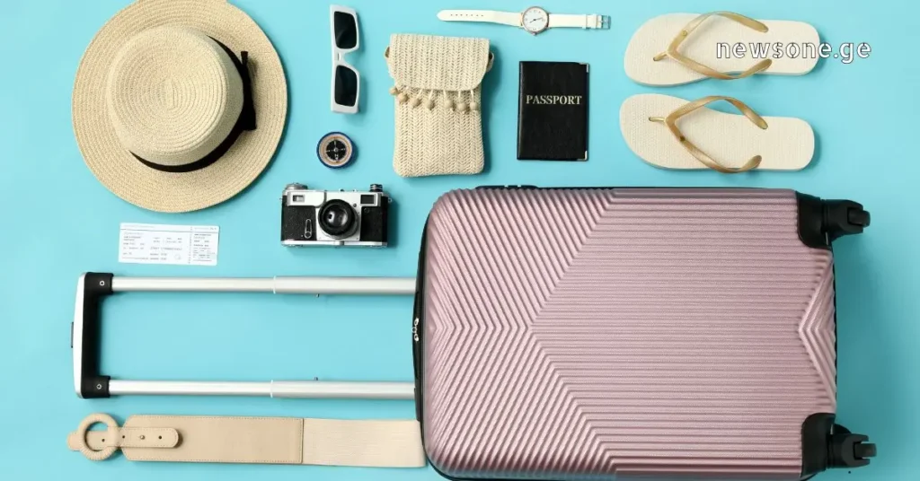 Flat lay of a pink travel suitcase with summer vacation essentials like a hat, passport, and camera on a blue background.