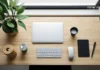 კომფორტი, როგორც წარმატების ფორმულა: რატომ არის გარემო პროდუქტიულობის საწინდარი? Top view of a minimalist wooden desk with a laptop, wireless keyboard, mouse, notebook, and a green plant near a window.