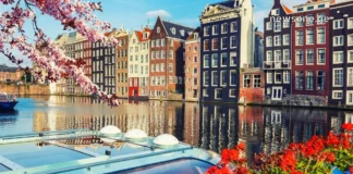 ამსტერდამი: თავისუფლების, არხების და ტიტების ქალაქი – სრული გზამკვლევი ქართველი მოგზაურებისთვის Colorful traditional canal houses in Amsterdam with spring cherry blossoms and a boat in the foreground.